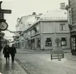Västerås. Köpmangatan mot norr med korsningen Hantverkargatan.
Åkerboms ur hänger utanför butik, Guldbutik, Didners, blomsteraffär m.fl. 1963.