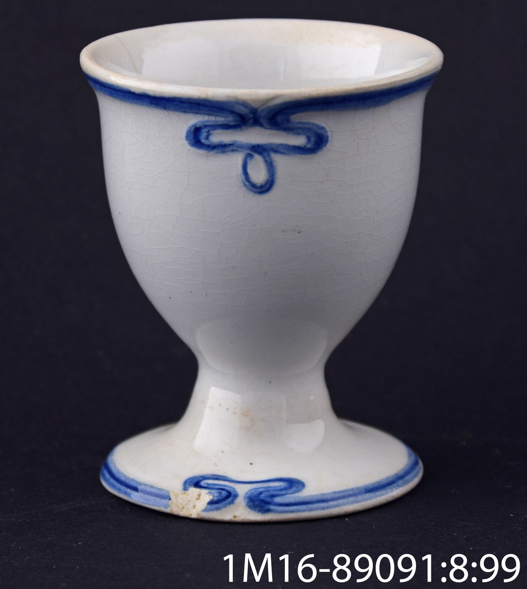 Äggkopp, i jugendstil med reliefdekor och handmålad mörkblå dekor. Tillverkad av Villeroy & Boch, Tyskland. Modellnamn: Pommern. Dekornamn: blaue Olga. Blå Olga. Dekornummer: 2069.