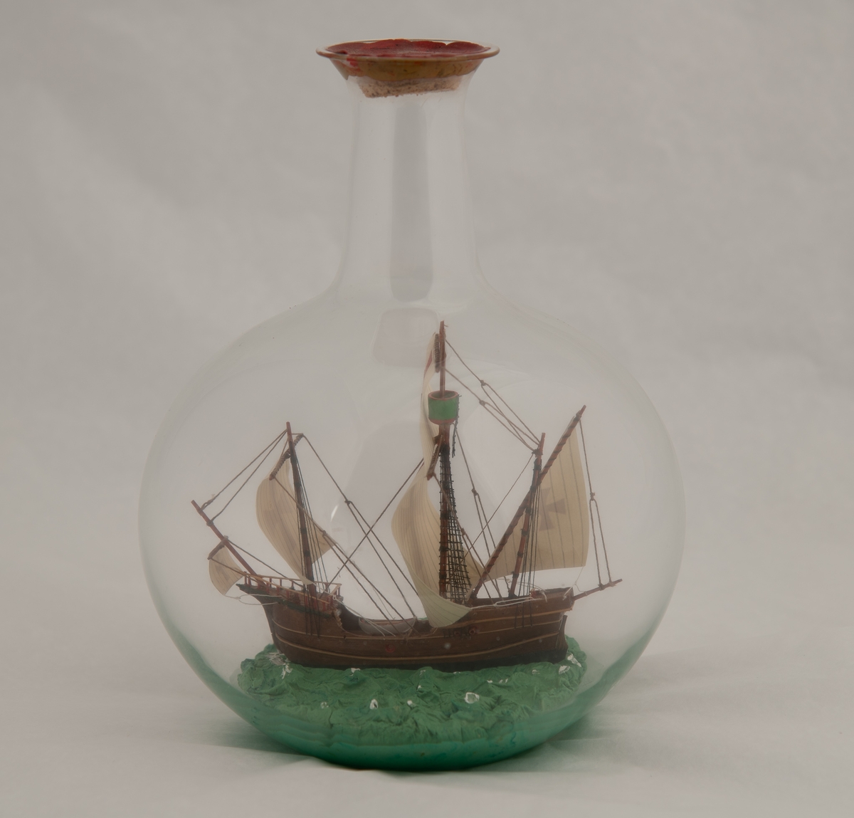 Flaskeskute i kuleformet flaske. Skuta er: "Santa Maria" , 1492 , en karakk , som var flaggskipet til Columbus.  Brunt skrog med lyse striper , seil med malteserkors i rødt.