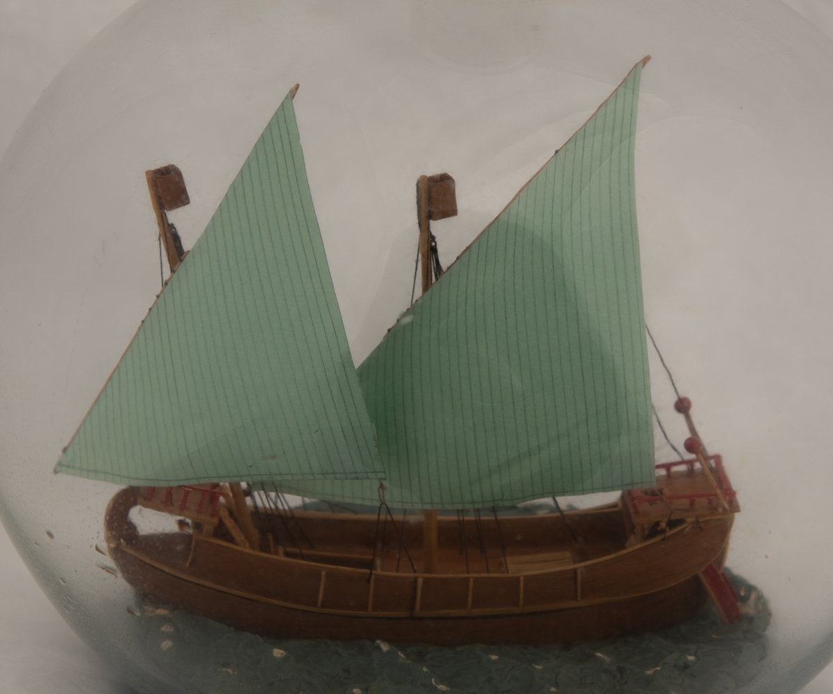 Flaskeskute; Veneziansk handelsfartøy , ca. 1200.  Kuleformet flaske i klart glass.  Brunt skrog med lysere belistning , lysegrønne seil (2 råseil) , rødt på akteroppbygningen.