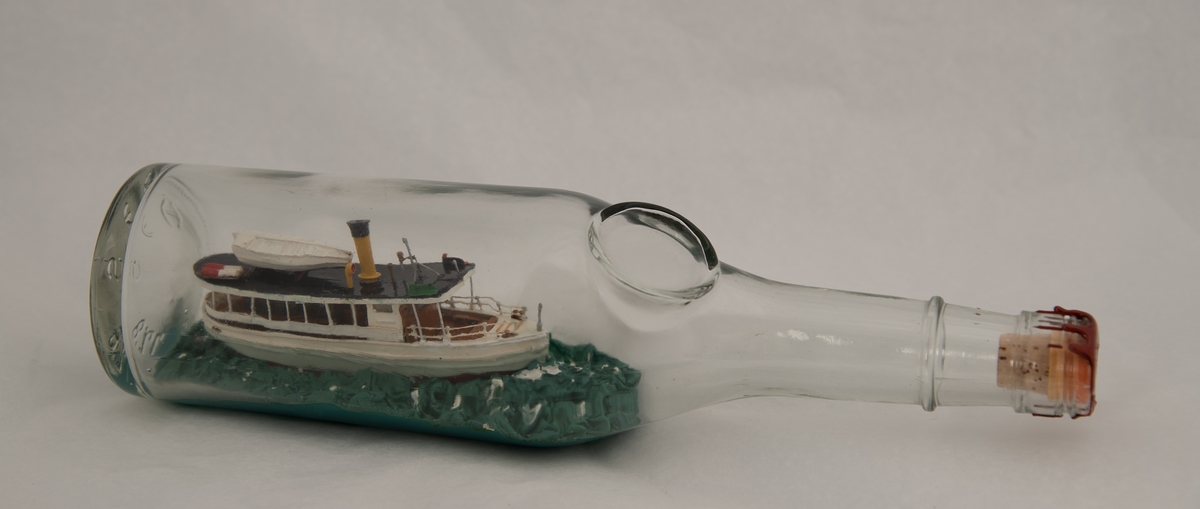 Flaskeskute: "Pelle" av Arendal.  Glass, svakt blågrønt, lukket med røft lakk. Flaskeskute bygget av  tre, bambus, knappenåler, og malt. Vinduer av plast. D/S "Pelle" hvitmalt med sort overdekk, gul skorstein med sort øverst, hvit livbåt, to hvite og røde nødraketter. Brunlakket passasjerdekk. Grønnblå, toppet sjø. Flaske med sirkulært merke for etikett på skulderen. En ring om halsen, har hatt skrukork: gjenger øverst. Innstemplet i flaskebunnen:  70 CL,