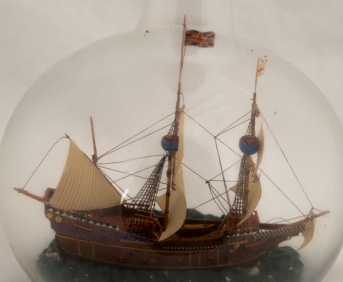 Flaskeskute; "Mayflower", en tremastet galleon fra 1615 , tok i l620 102 utvandrere fra England til Amerika. Symbol for frihetsrettigheter og religiøs toleranse.  Kuleformet glassflaske. Brunt skrog , dekorert i akterspeilet med border i blått , rødt , sort , hvitt og gult. Britisk flagg.