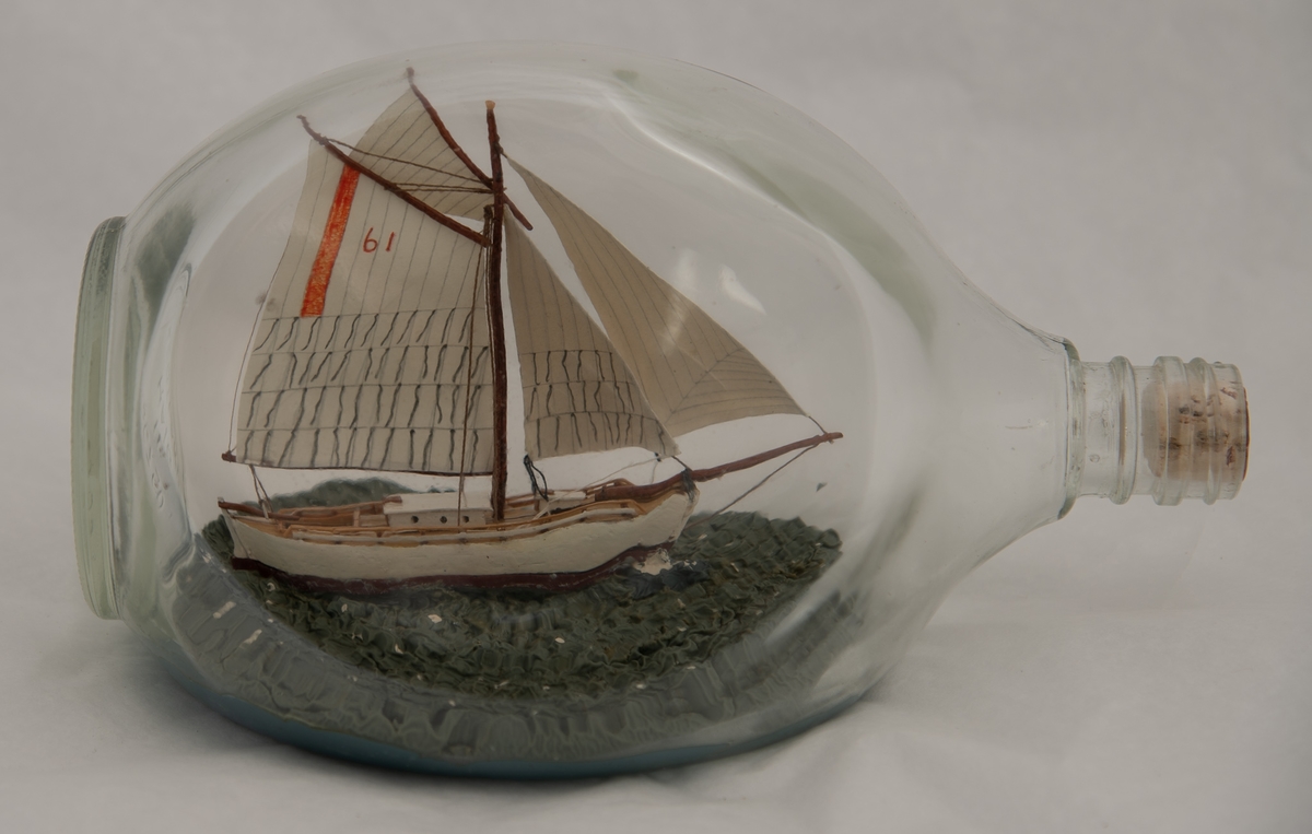 Flaskeskute: "Krabben" , losbåt nr. l (Colin Archer)  Flaske i klart glass med trekantet form.  Hvitmalt skrog , brun under vannlinjen. Seil med oransje stripe og tallet 61 i oransje. Blågrønn sjø.