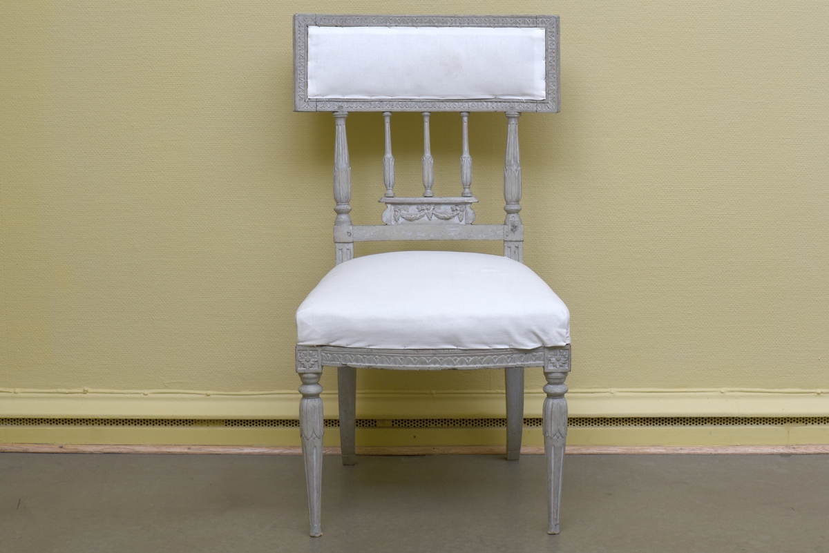 Gråvit stol i gustaviansk stil av björk på runda, avsmalnande ben. Benens övre del är dekorerade med bladkrans. Den främre sargen är något svängd och dekorerad med en äggstavslist. Den rektangulära ryggbrickan bärs upp av totalt fem, tre mindre och två större, ståndare. Ryggbrickans ramvirke är dekorerad med äggstavslist. Stolen är provisoriskt klädd på sits och i rygg med ett vitt bomullstyg.