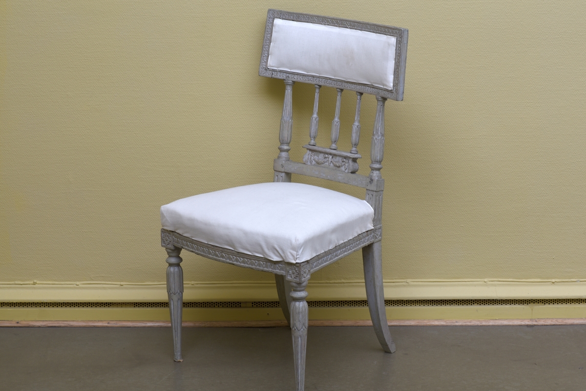 Gråvit stol i gustaviansk stil av björk på runda, avsmalnande ben. Benens övre del är dekorerade med bladkrans. Den främre sargen är något svängd och dekorerad med en äggstavslist. Den rektangulära ryggbrickan bärs upp av totalt fem, tre mindre och två större, ståndare. Ryggbrickans ramvirke är dekorerad med äggstavslist. Stolen är provisoriskt klädd på sits och i rygg med ett vitt bomullstyg.