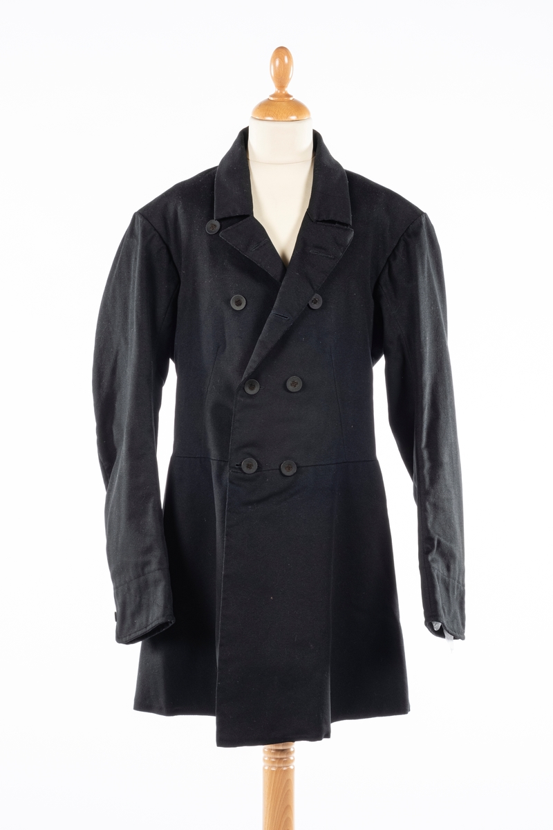 Dobbelt spent frakk som kneppes med fire knapper på hver side. De øverste kan ikke kneppes. Knappene er kledd med silke med fløyelsornament i midten. Hempe i nakkelinning. Det er splitt med legg fra livet og ned, med en knapp over splitten på hver side. Det er også to knapper lenger ned i splitten, og en knapp på hver mansjett. Frakken er foret.