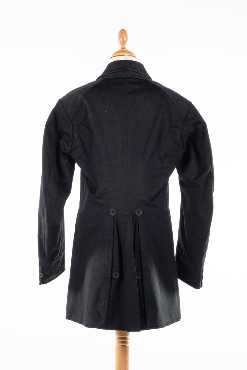 Dobbelt spent frakk som kneppes med fire knapper på hver side. De øverste kan ikke kneppes. Knappene er kledd med silke med fløyelsornament i midten. Hempe i nakkelinning. Det er splitt med legg fra livet og ned, med en knapp over splitten på hver side. Det er også to knapper lenger ned i splitten, og en knapp på hver mansjett. Frakken er foret.