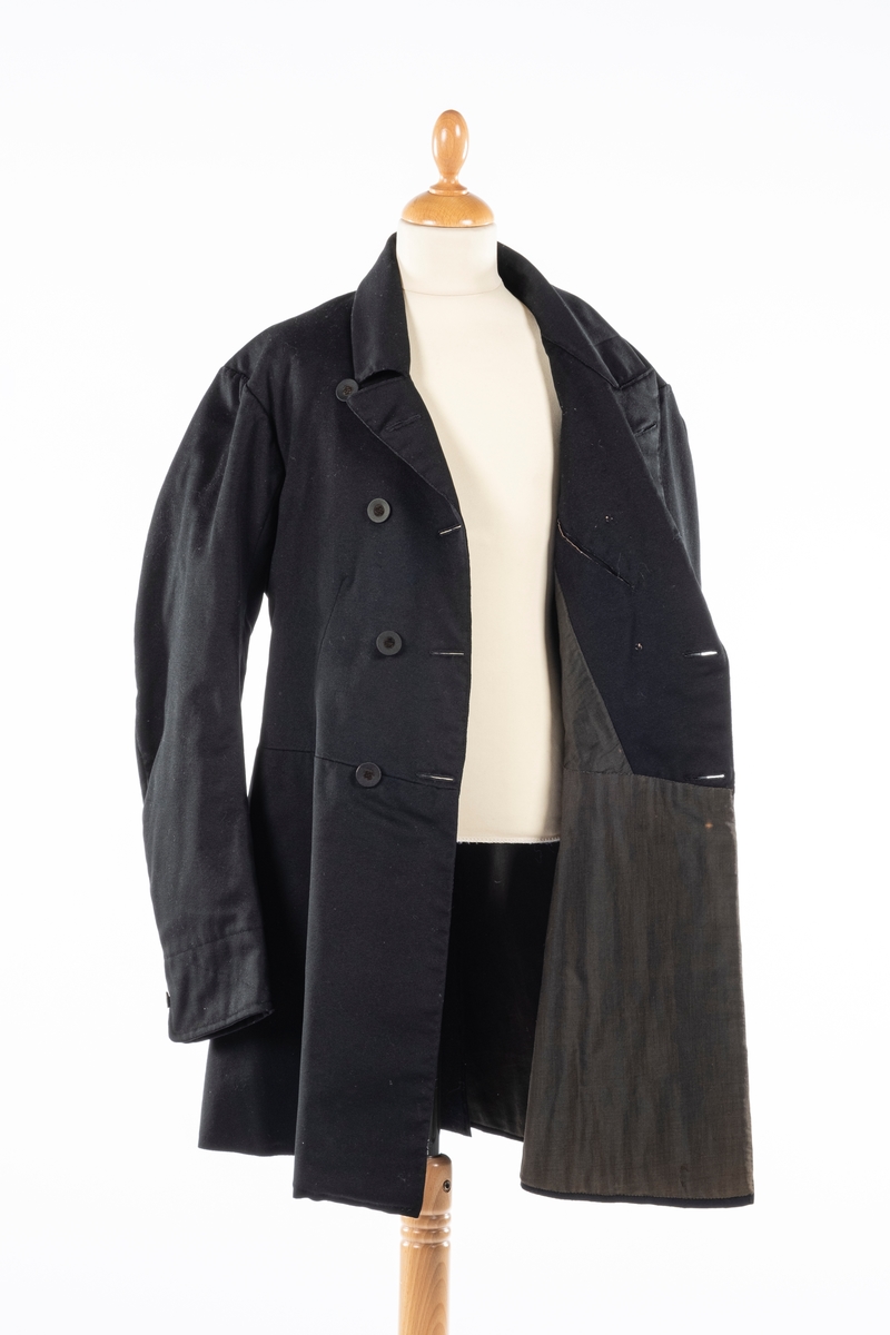 Dobbelt spent frakk som kneppes med fire knapper på hver side. De øverste kan ikke kneppes. Knappene er kledd med silke med fløyelsornament i midten. Hempe i nakkelinning. Det er splitt med legg fra livet og ned, med en knapp over splitten på hver side. Det er også to knapper lenger ned i splitten, og en knapp på hver mansjett. Frakken er foret.