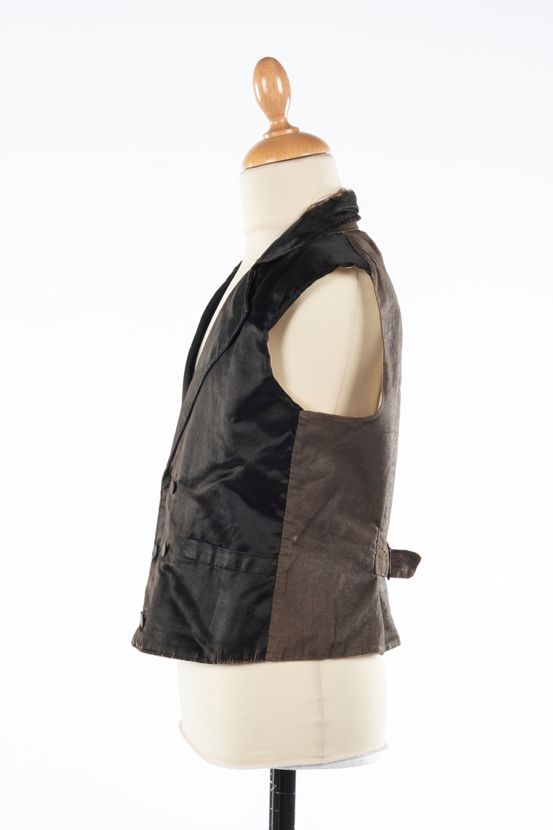 Vest med forstykke i silke og bakstykke i bomull. Den er foret med lin. Det er dobbelspent knepping og spensel i rygg. Knappene er trukket med silke.