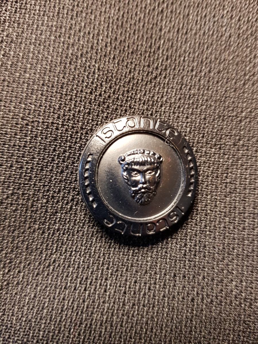 Hoftelang, dobbeltspent, med 6 metallknapper, med Versace-husets "Istante"-logo - Zevs hode - i relieff. Lange slag, og kun de nederste 4 knapper skal kneppes. Lapellene (slagene) er spisse og lengre enn kragen. Ett ekstra knapphull for innvendig knapping, men knappen på innsiden av venstre forstykke mangler. To-søms ermer med falsk splitt, lukket knapphull og pynteknapp. Horisontal lomme med belegg på hver hofte og stikklomme med belegg på venstre bryst. Innvendig, passepoilert lomme på hvert bryst, den høyre lukket med klaff og usynlig knapp. Søm midt bak og splitt nederst i hver panelsøm bak. Forstykkene er skåret rettvinklet i nederkant midt foran. Helfôret med svart fôrsilke. Kragestanden og kragen er kledd med filtaktig materiale på baksiden og jakken har puter i skuldrene.