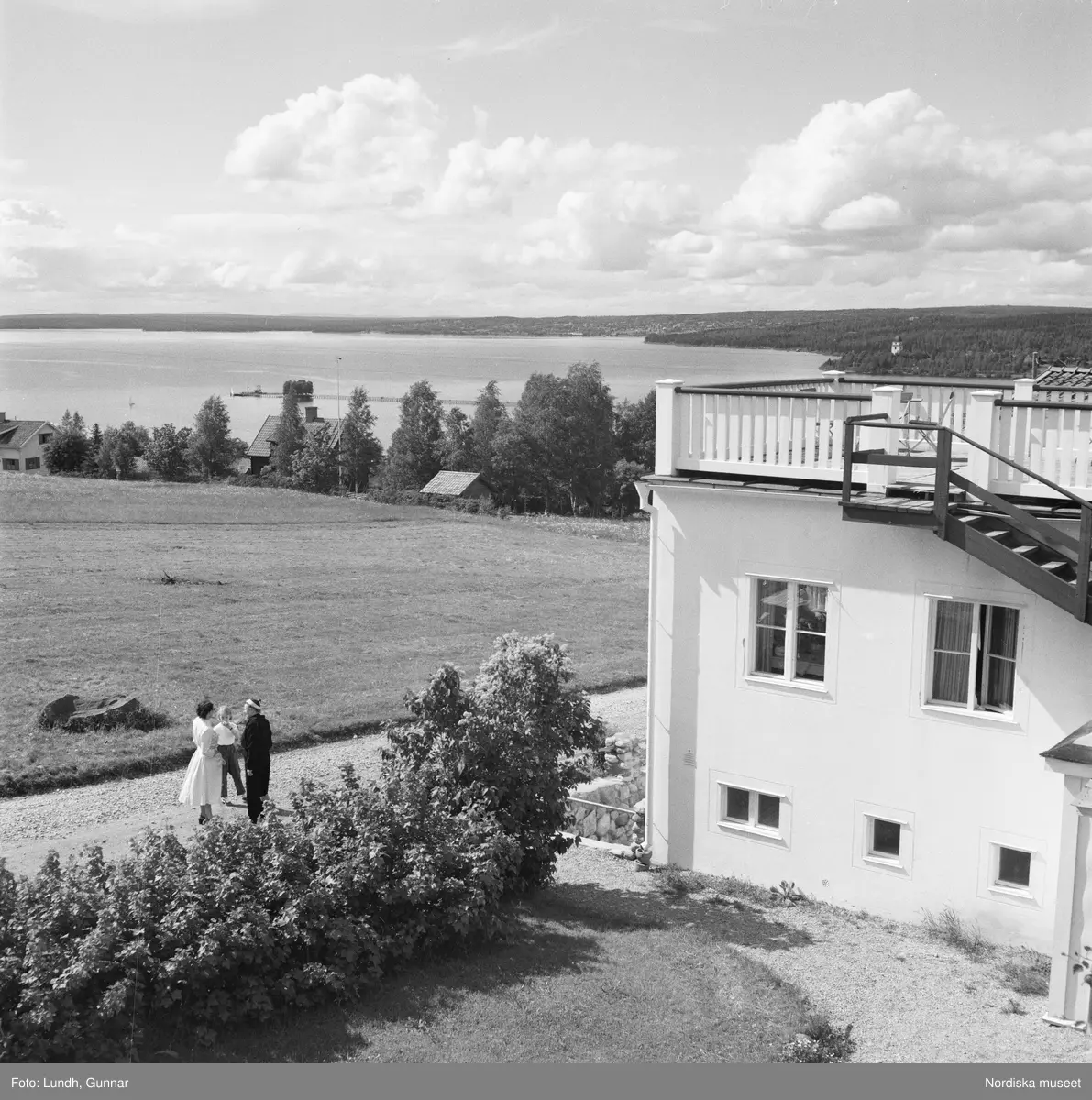 Motiv: Rättvik ;
Exteriör av hus, interiör med utsikt genom fönster med flaggstänger med nationsflaggor, tre kvinnor sitter vid ett trädgårdsbord vid parkerade bilar, porträtt av en man som sitter på en förstukvist, en kvinna och tre män spelar boccia.