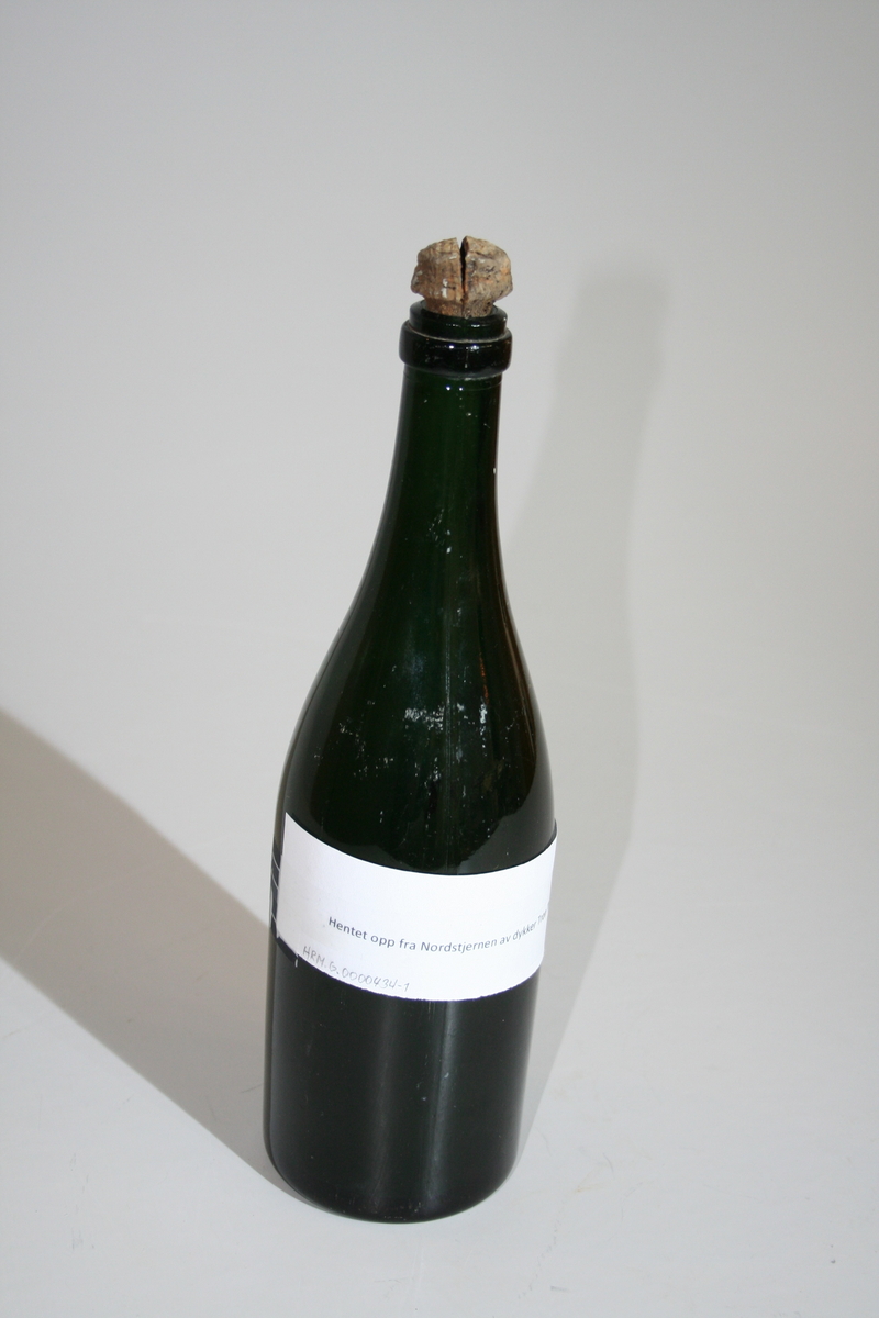 Tom flaske i farget glass og med opphøyet bunn. Korken sitter fortsatt på og har en "champagnekork" fasong. Korken har en sprekk i tioppen. Et papir sitter tapet fast med informasjon om giver