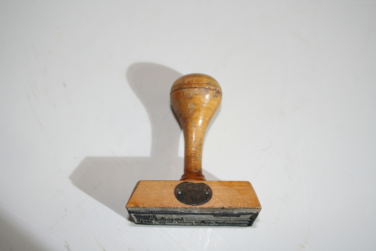 Stempel i tre med retangulær stempelflate. Dreid skaft. Et ovalt lite metallskilt på siden med produsentlogo. Lakkert