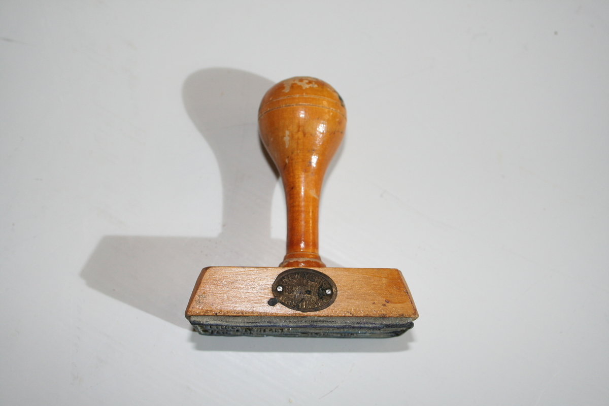 Stempel i tre med retangulær stempelflate. Dreid skaft. Et ovalt lite metallskilt på siden med produsentlogo. Lakkert