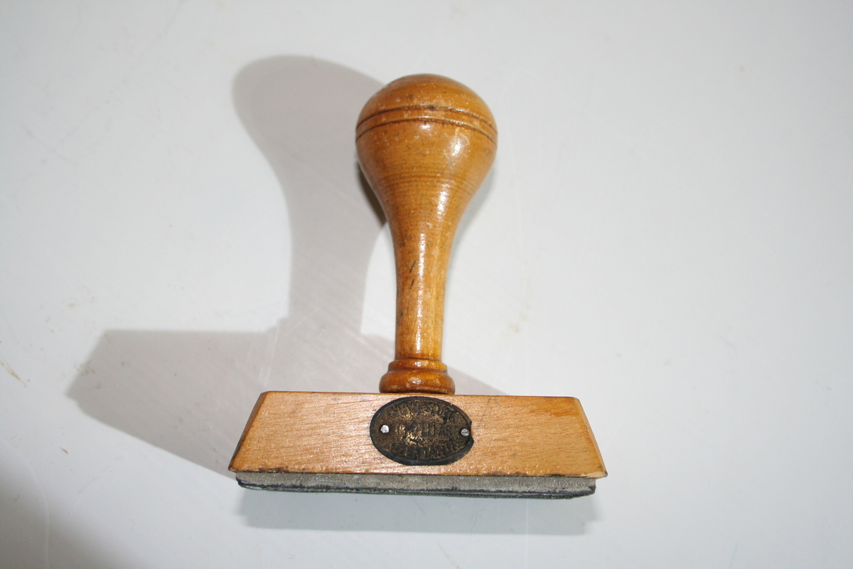 Stempel i tre med retangulær stempelflate. Dreid skaft. Et ovalt lite metallskilt på siden med produsentlogo. Lakkert