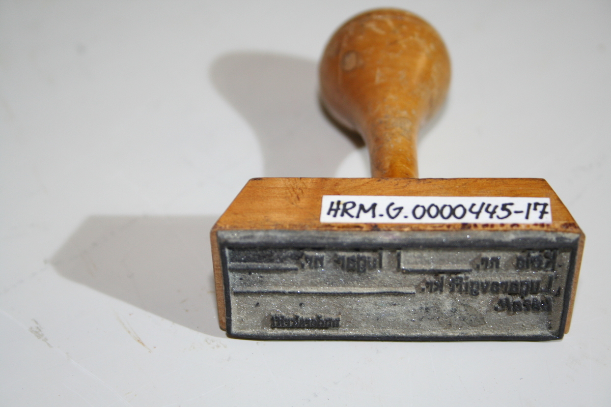 Stempel i tre med retangulær stempelflate. Dreid skaft. Et ovalt lite metallskilt på siden med produsentlogo. Lakkert