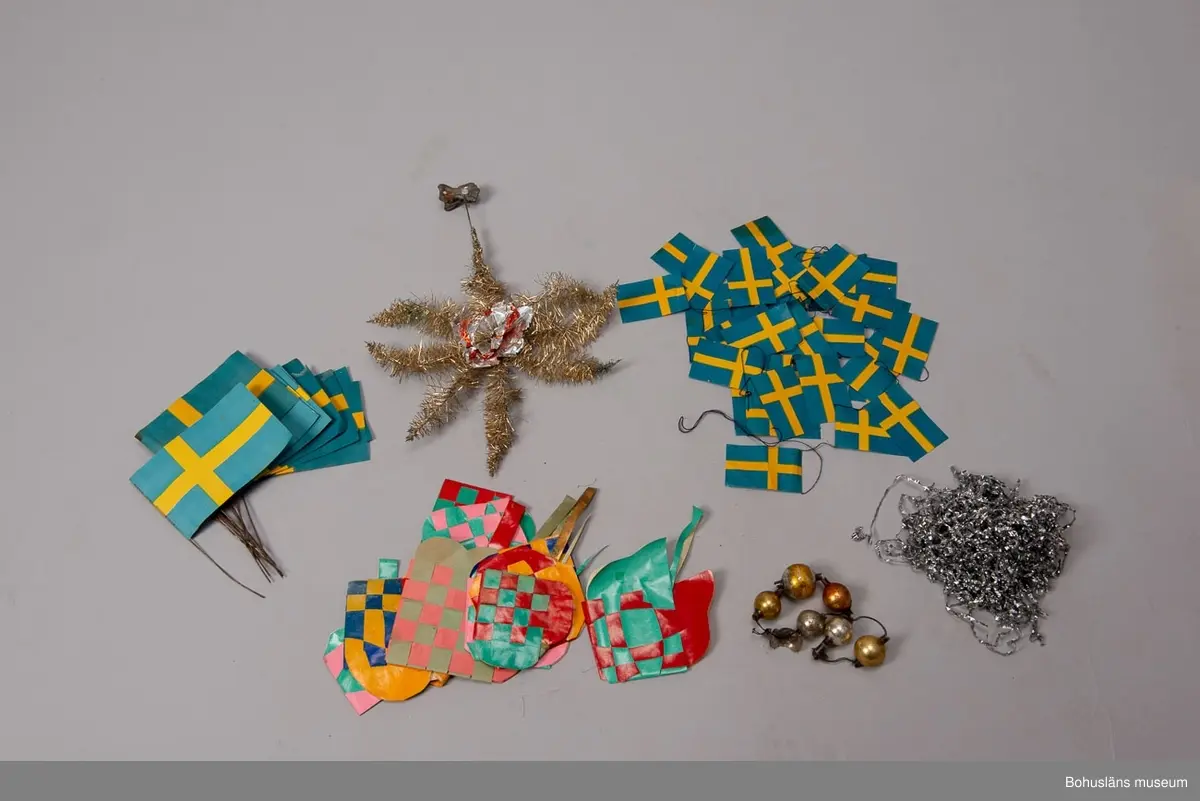 Låda av brun papp. På locket påklistrad,delvis bortsliten etikett med text: "Herman Radyn Uddevalla". Lådan innehåller: 19 st. julgransljushållare, glitter i silver, glitterband i guld, 7 st. julgranskarameller av papper, 1 toppstjärna, 12 st. hjärtan av papper i olika färger, 6 st. små glasbollar i sytråd, svenska flaggor i tråd, 12 st. svenska flaggor med järntråd,