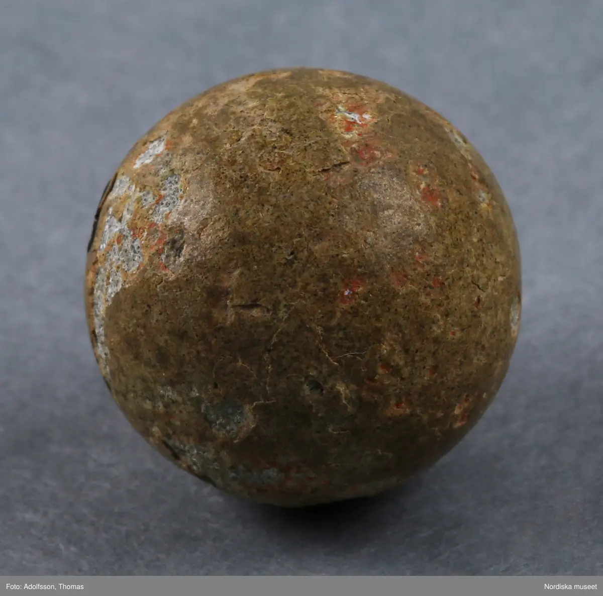 Kula av sten, ljust gråbrun. Den ursprungligen blanka ytan har många små slagskador, kulan är väl använd. Den låg i den vänstra nedre fickan på en mycket väl använd, sliten kavaj använd av en pojke, ca 7-8 år gammal, omkring 1910. 
/Helena Lindroth 2019-02-14