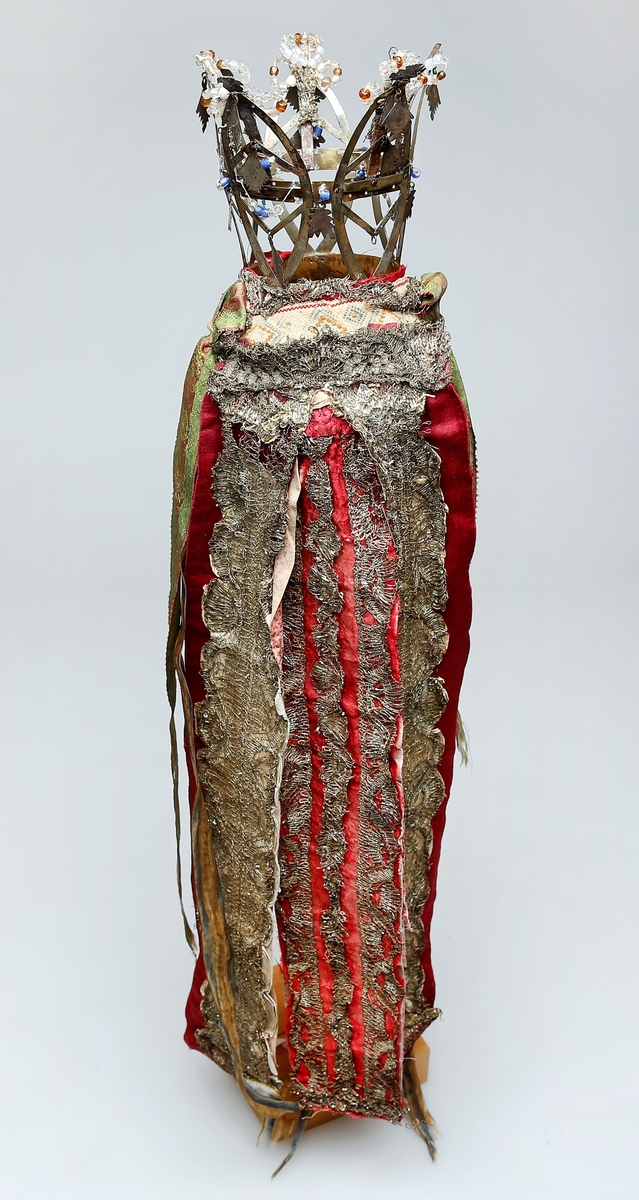 Brudekrone i metall med fem spir. Krona er pyntet med metallheng og glassperler. Det er påsydd valk nederst, med brodert bånd og metallknipling rundt valken.

Bak på det broderte båndet rundt valken, er det festet bånd. På midten er det et bredt bånd i rødt med metallkniplinger, med brede bånd med lys bunn og med en rød kant vendt ut fra den røde midtbåndet. Metallkniplinger er festet til de tre brede båndene. Deretter fire smalere bånd på hver side av de brede båndene. Disse er festet samlet med en liten sløyfe på det bredeste der det er festet på valken. 

En plastkam er påsydd valken sammen med to hvite, nyere bånd. Antagelig gjort i nyere tid for å feste krona og knyte under haka.