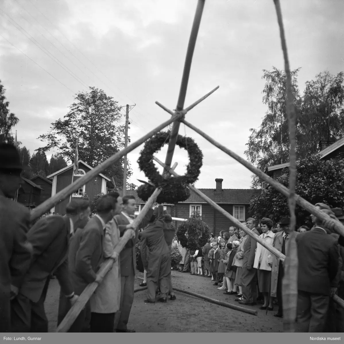 Motiv: Tibble midsommar ;
Kvinnor och män arbetar med att pryda en midsommarstång inför en publik, resning av midsommarstång.