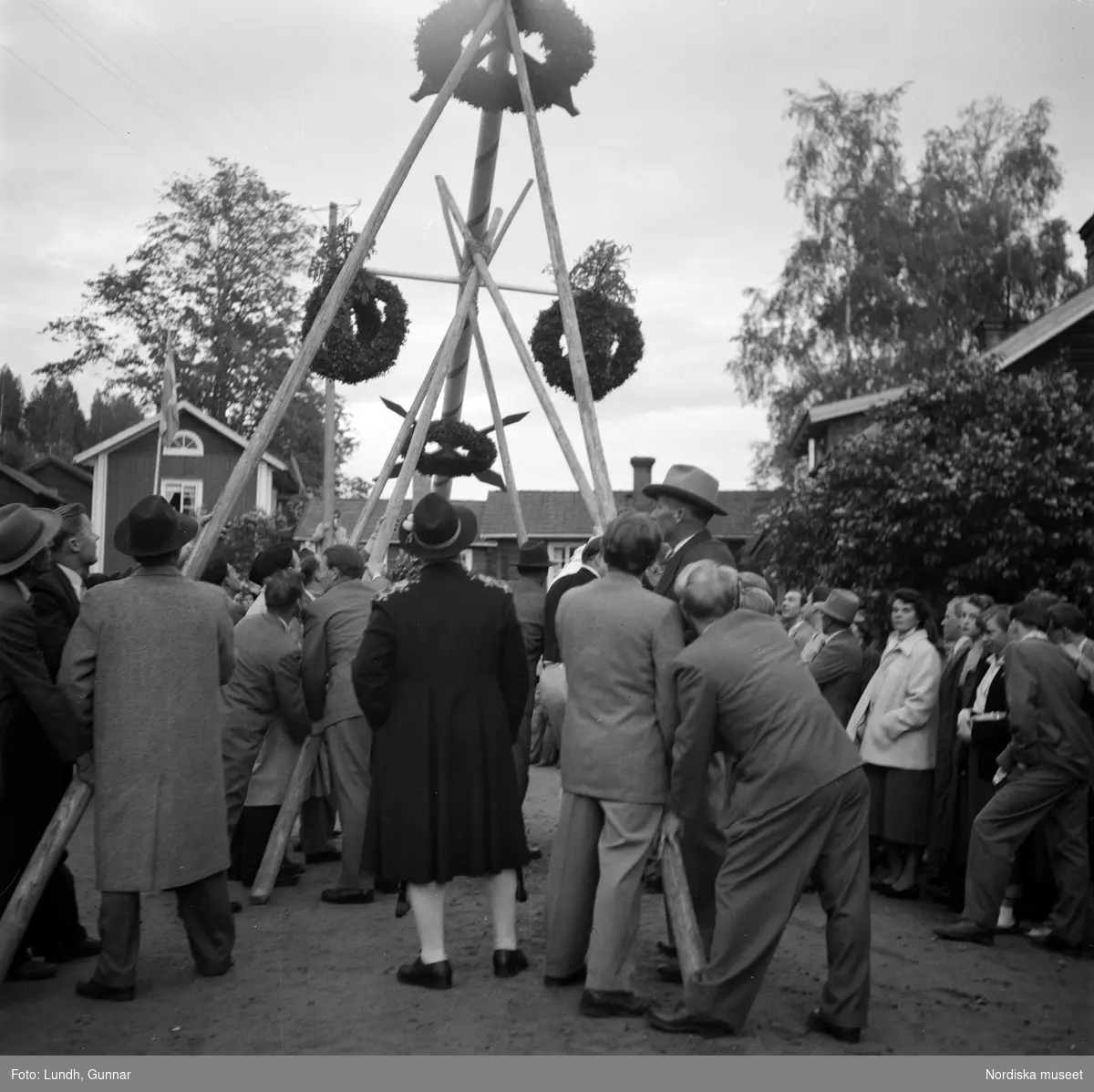 Motiv: Tibble midsommar ;
Kvinnor och män arbetar med att pryda en midsommarstång inför en publik, resning av midsommarstång.