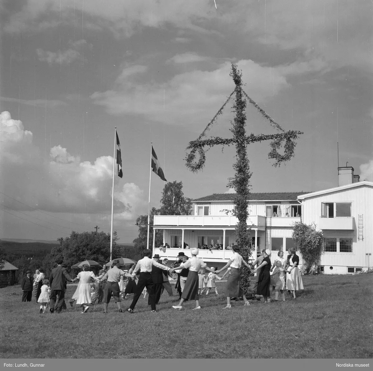 Motiv: Lerdalshöjden ;
Porträtt av en flicka som håller blommor, vuxna och barn dansar runt en midsommarstång, tre män i folkdräkt spelar fiol vid en midsommarstång.