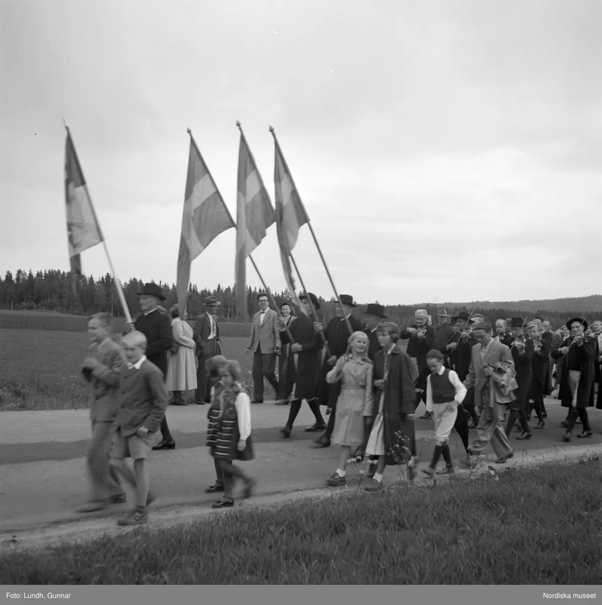 Motiv: (ingen anteckning) ;
Porträtt av en kvinna i folkdräkt och en man som sitter på marken, män och kvinnor i folkdräkt går i ett proccesionståg med flaggor och kvinnor bär en lövad girlang, människor åker på en hästdragen vagn, en folksamling går i ett processionståg.