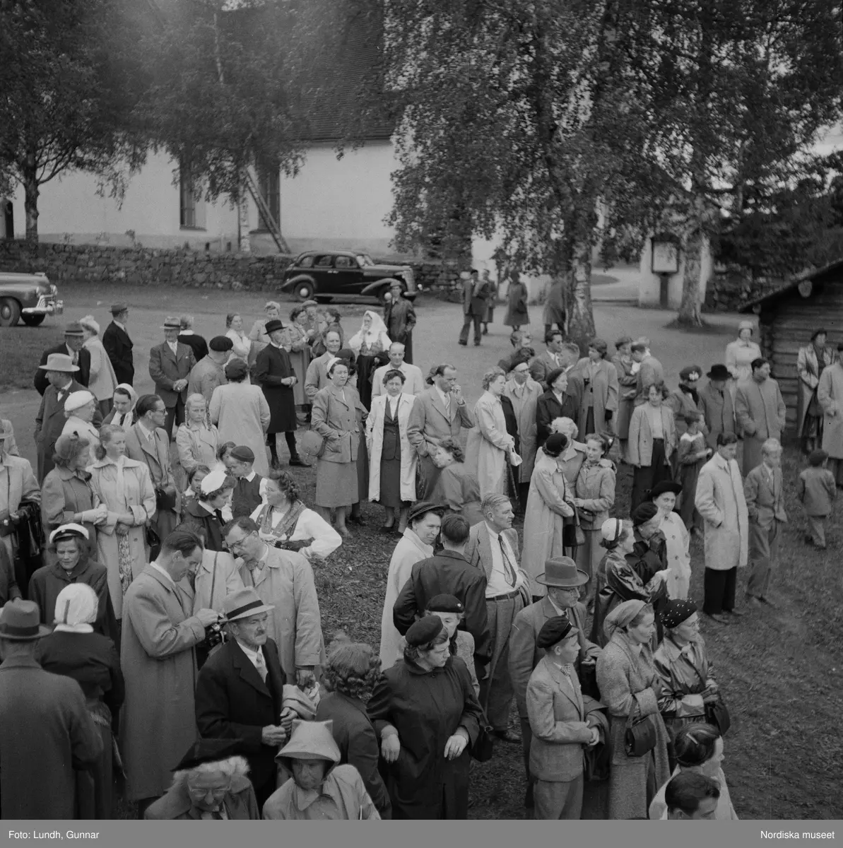 Motiv: (ingen anteckning) ;
En folksamling vid midsommarfirande, en folksamling står på stranden vid en sjö, kvinnor och män i folkdräkt ror en kyrkbåt.