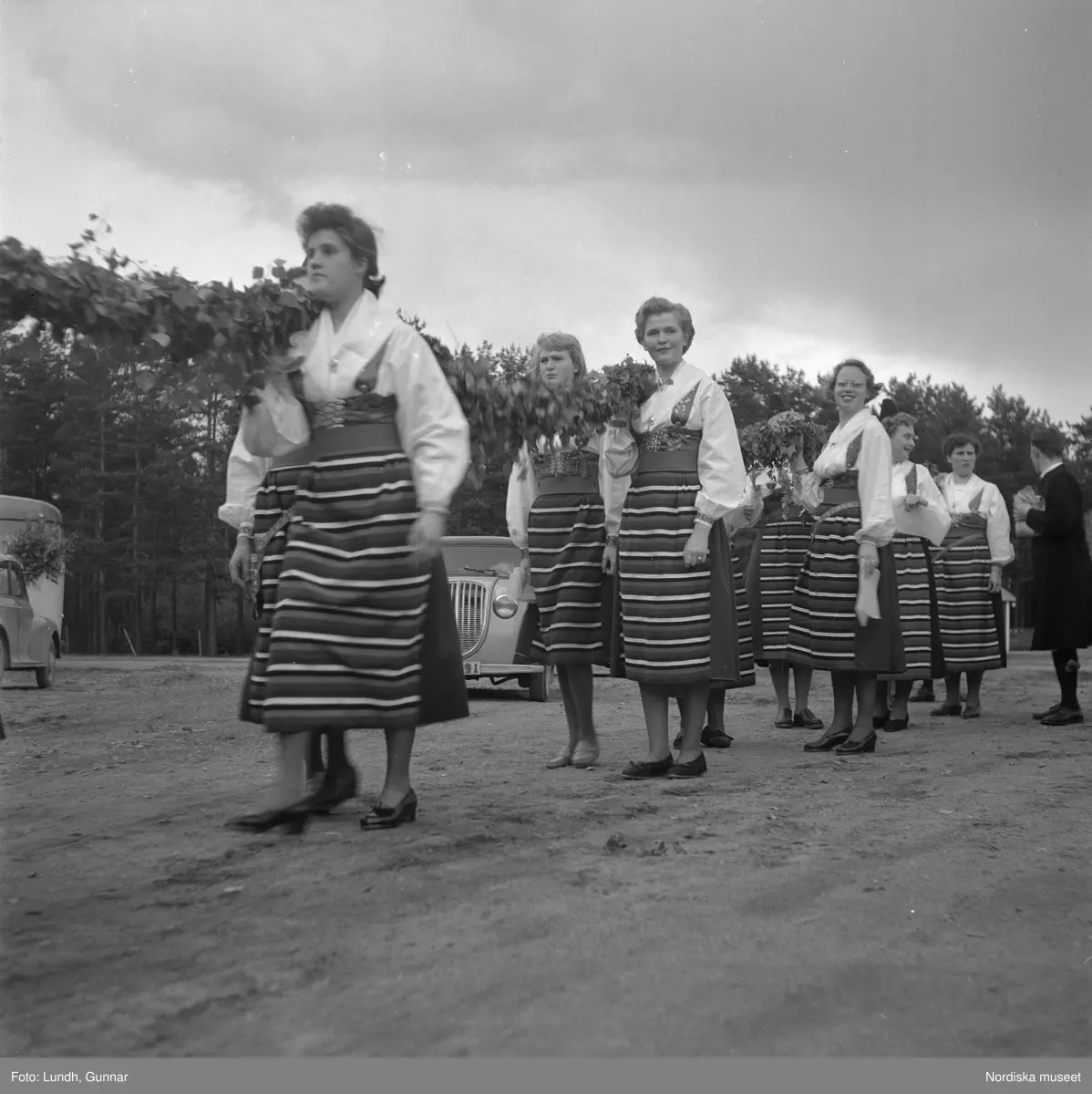 Motiv: midsommarfirande i Rättvik - Leksand ;
En pojke i folkdräkt och en flicka står vid en midsommarstång, en flicka pussar en pojke, en kvinna och en man handlar trasmattor av en man i en försäljningsvagn med text "Hemslöjd", en kvinna står vid en bil med kopparsaker till försäljning och en skylt "Ruben I Redin", en kvinna och en man i folkdräkt håller lövade ringar, kvinnor i folkdräkt går på en väg, en man kör en hästdragen vagn, ett led av kvinnor och män i folkdräkt bär en lövad girlang, människor åker på hästdragna vagnar.