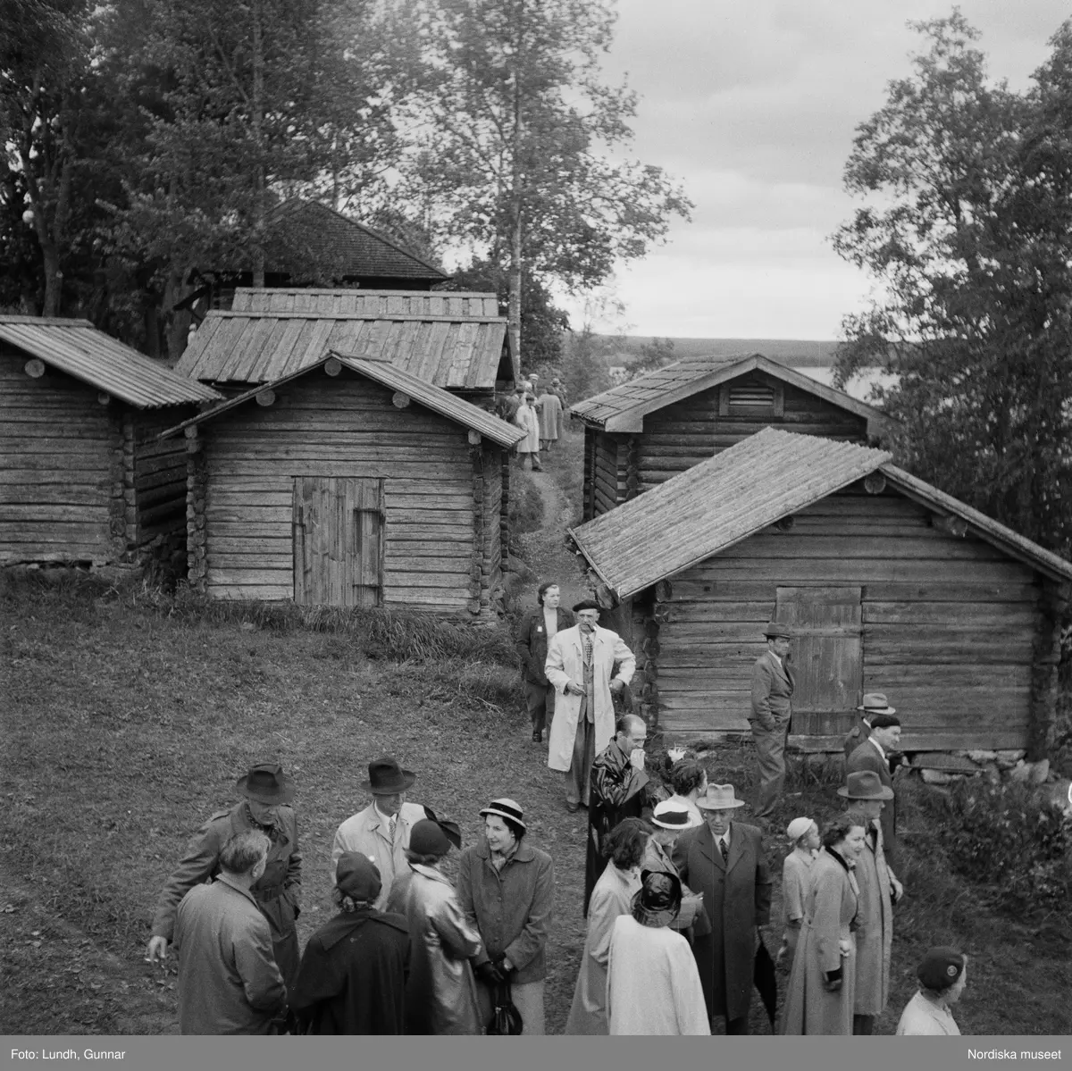 Motiv: (ingen anteckning) ;
En folksamling vid midsommarfirande, en folksamling står på stranden vid en sjö, kvinnor och män i folkdräkt ror en kyrkbåt.