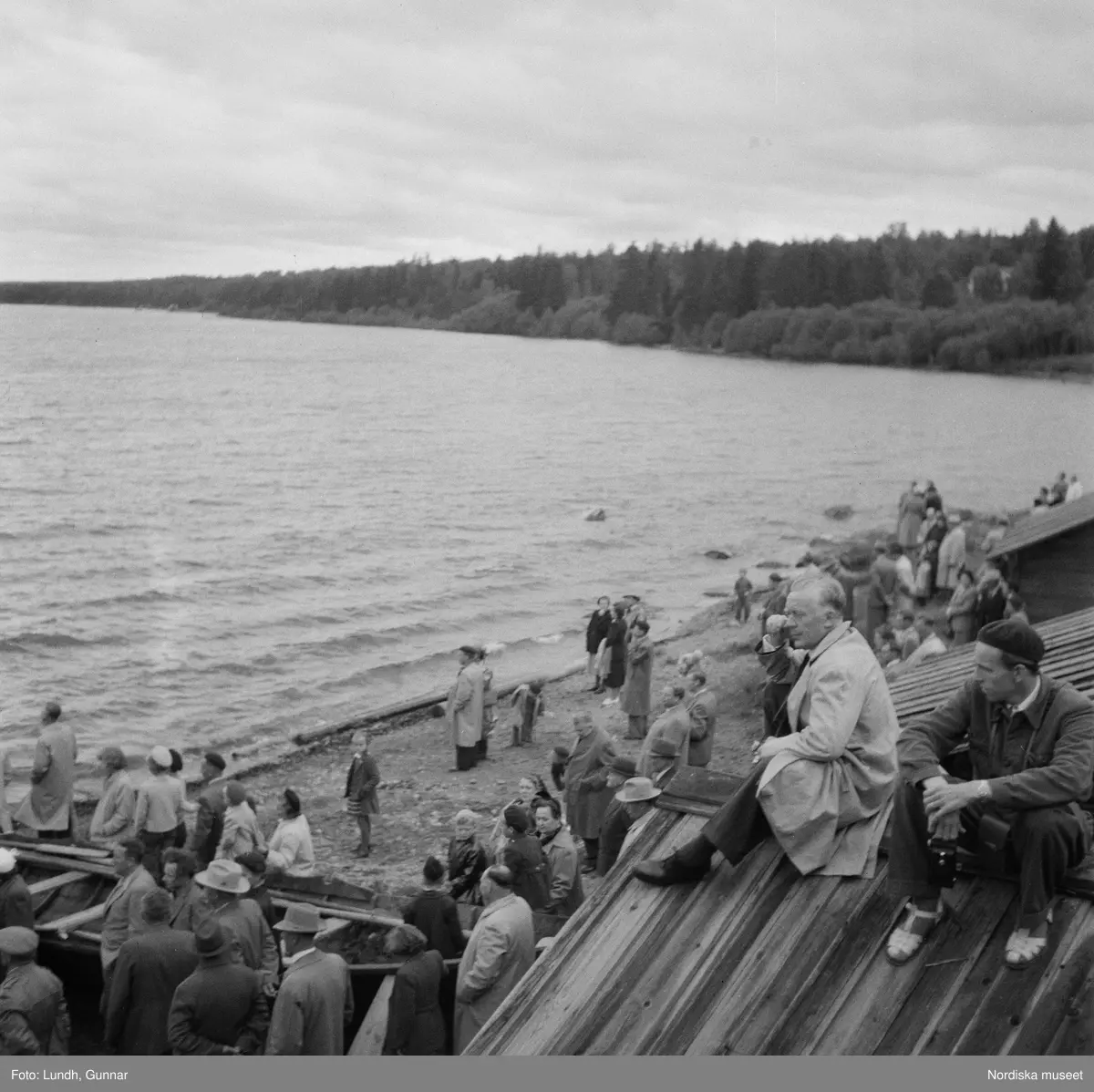 Motiv: (ingen anteckning) ;
En folksamling vid midsommarfirande, en folksamling står på stranden vid en sjö, kvinnor och män i folkdräkt ror en kyrkbåt.