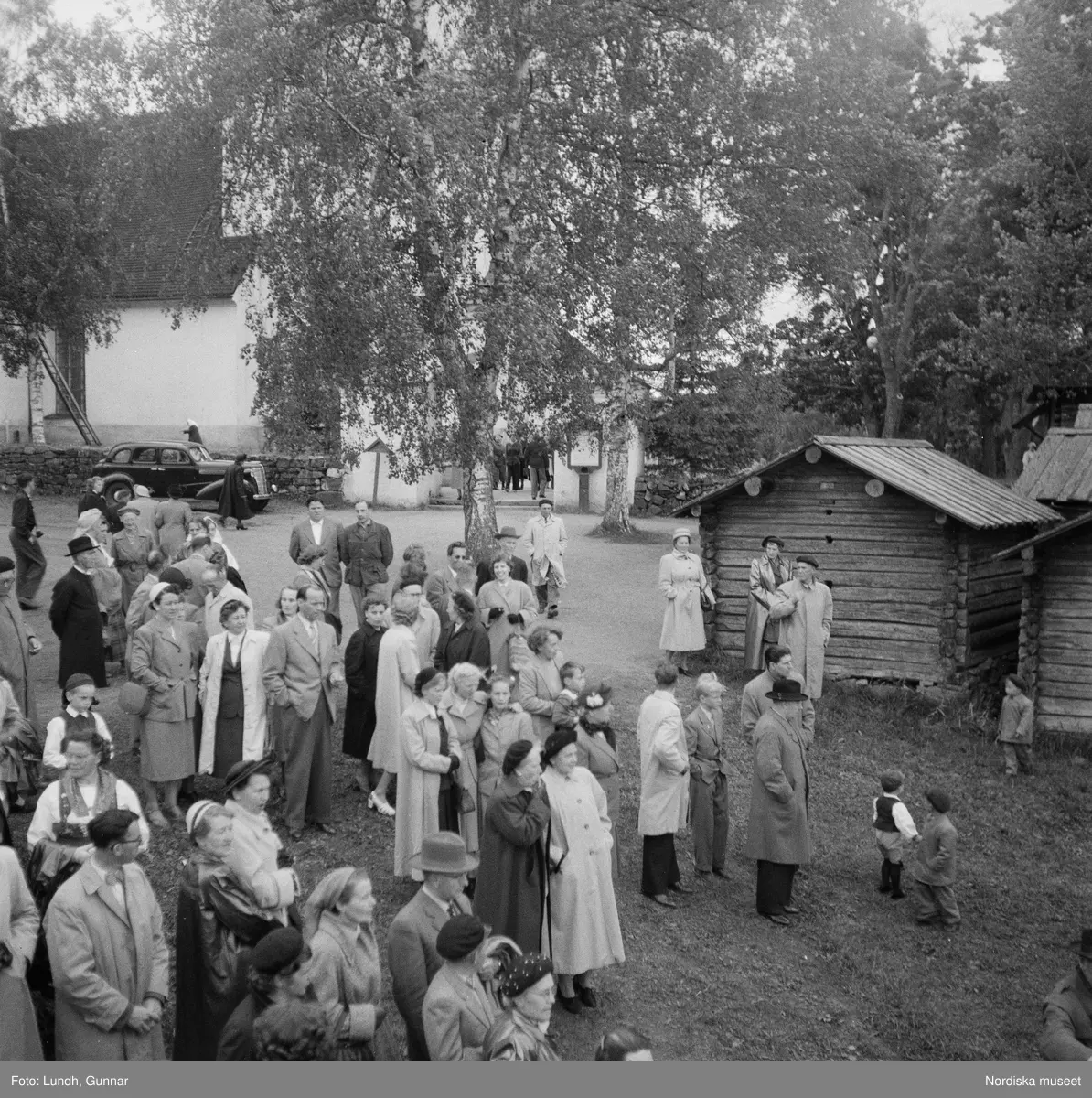Motiv: (ingen anteckning) ;
En folksamling vid midsommarfirande, en folksamling står på stranden vid en sjö, kvinnor och män i folkdräkt ror en kyrkbåt.