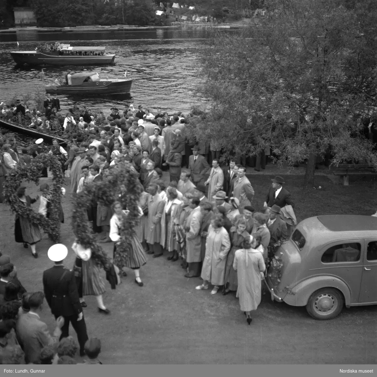 Motiv: (ingen anteckning) ;
En folksamling står kring en midsommarstång, en folksamling vid ett hus, en kyrkbåt ros av kvinnor och män i folkdräkt, en folksamling står vid en brygga med en kyrkbåt.