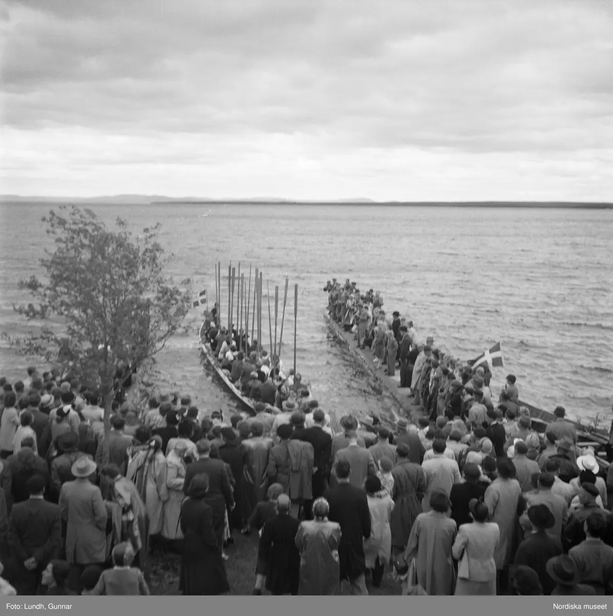 Motiv: (ingen anteckning) ;
En folksamling vid midsommarfirande, en folksamling står på stranden vid en sjö, kvinnor och män i folkdräkt ror en kyrkbåt.