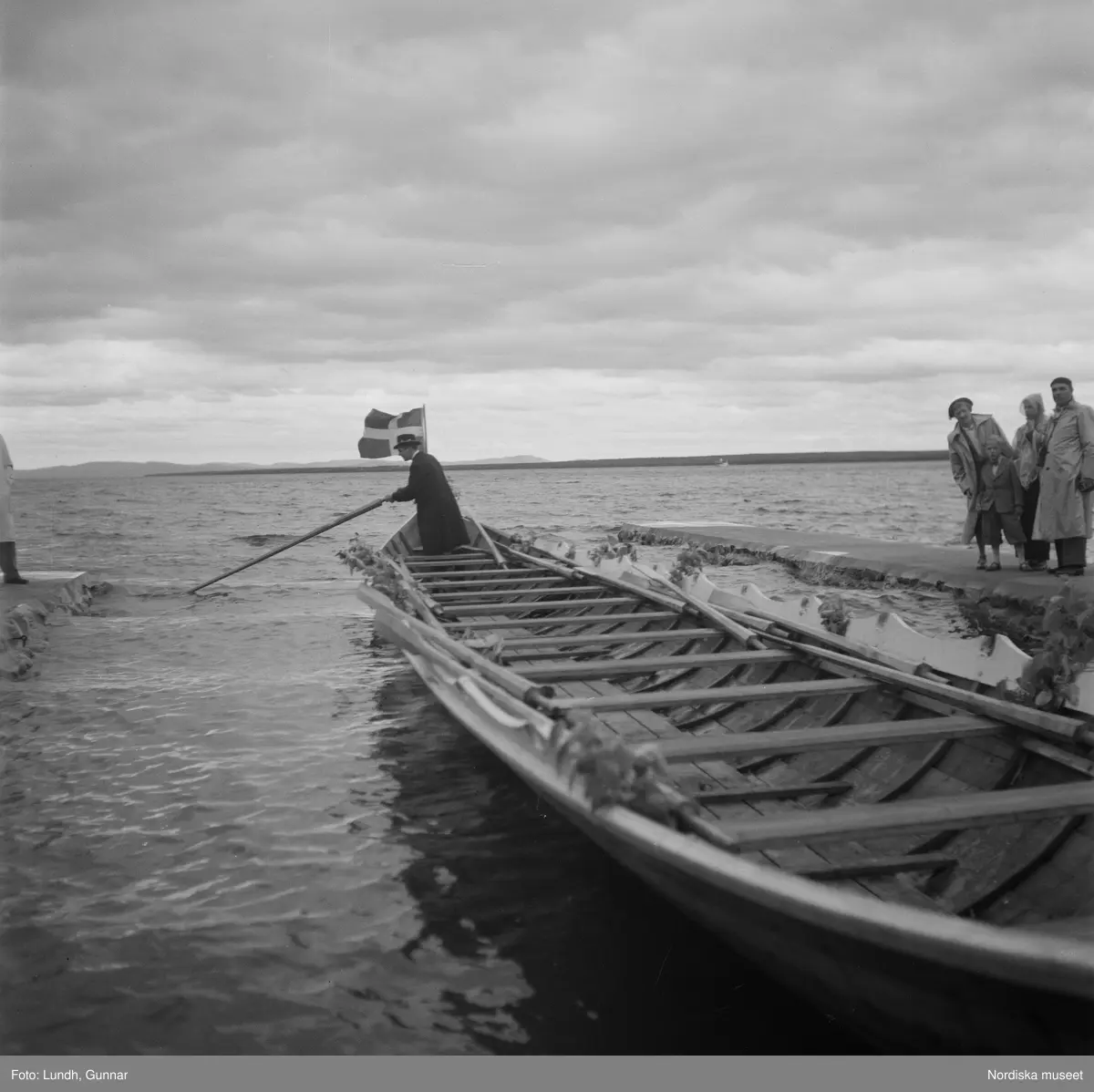 Motiv: (ingen anteckning) ;
En folksamling vid midsommarfirande, en folksamling står på stranden vid en sjö, kvinnor och män i folkdräkt ror en kyrkbåt.