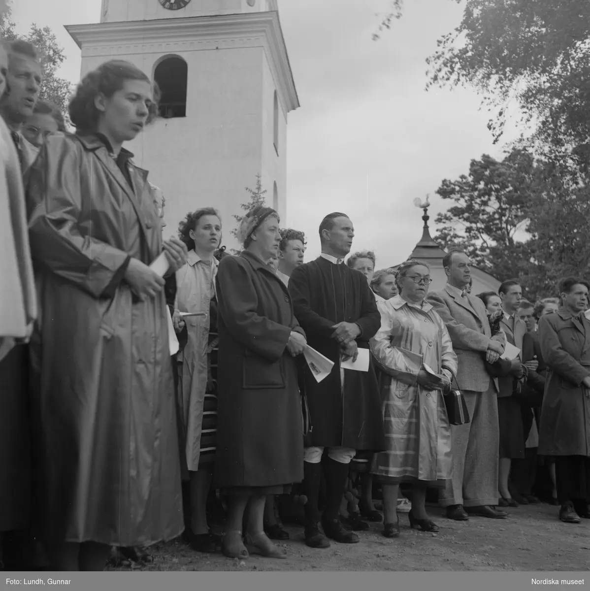 Motiv: (ingen anteckning) ;
En folksamling vid Rättviks kyrka, en folksamling står vid en brygga när kvinnor och män går ombord på två kyrkbåtar.