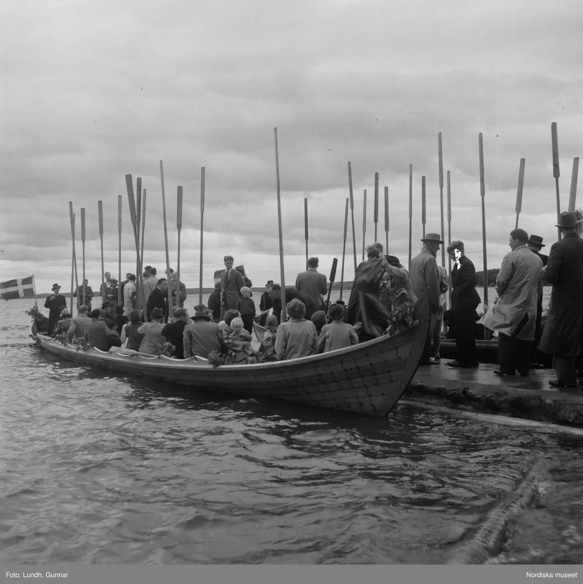 Motiv: (ingen anteckning) ;
En folksamling står vid en brygga med två kyrkbåtar, kvinnor och män ror kyrkbåtar i samband med midsommarfirande.