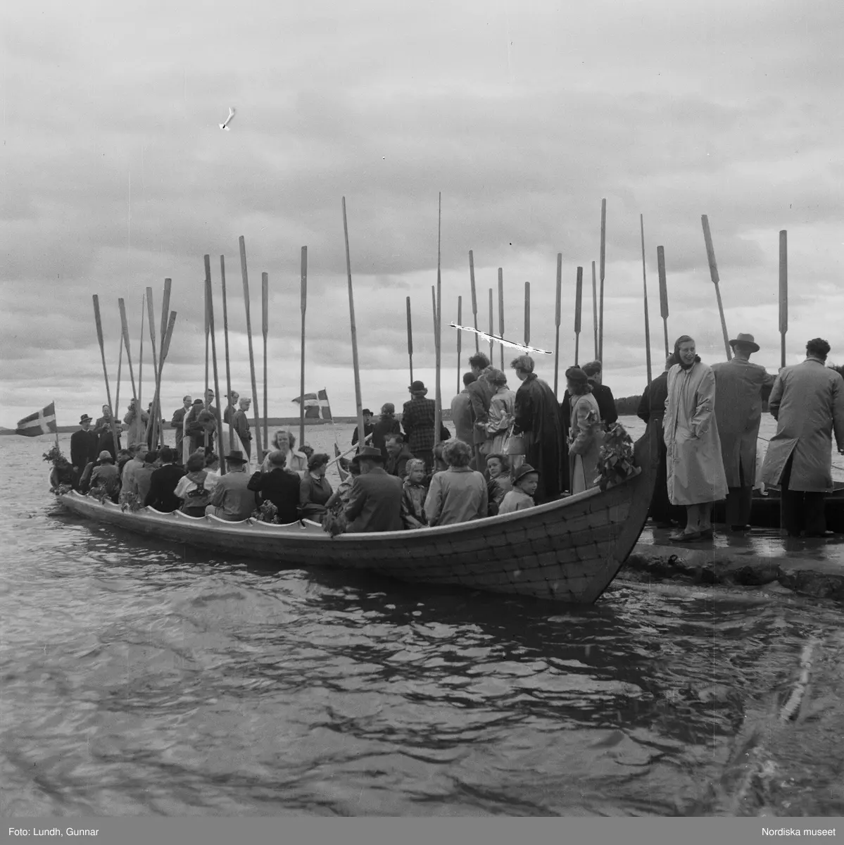Motiv: (ingen anteckning) ;
En folksamling står vid en brygga med två kyrkbåtar, kvinnor och män ror kyrkbåtar i samband med midsommarfirande.