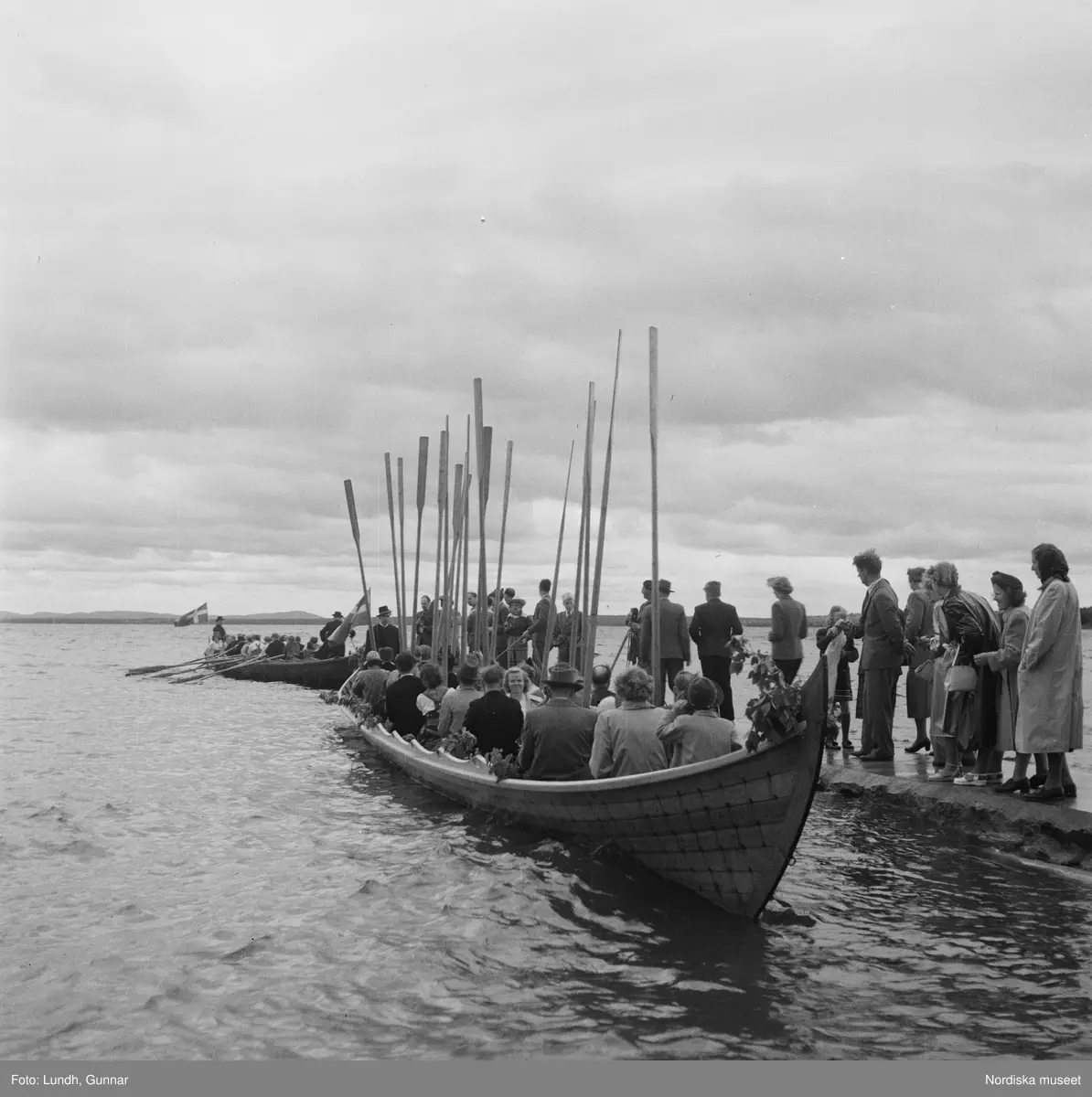 Motiv: (ingen anteckning) ;
En folksamling står vid en brygga med två kyrkbåtar, kvinnor och män ror kyrkbåtar i samband med midsommarfirande.