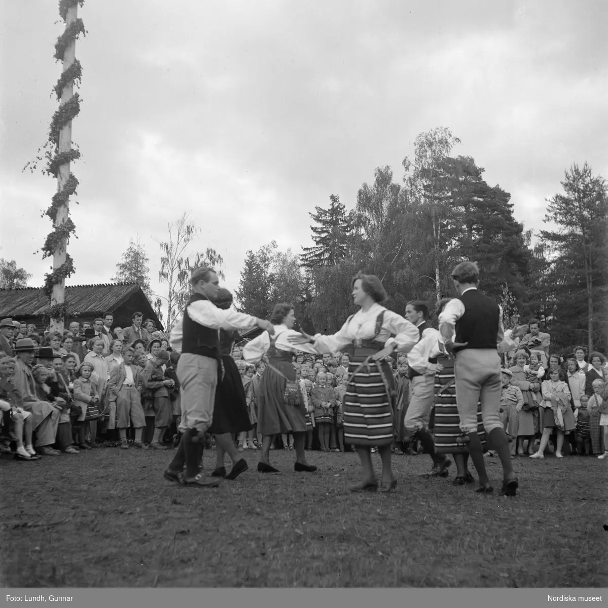 Motiv: (ingen anteckning) ;
Kvinnor och män i folkdräkt dansar vid en midsommarstång inför en publik vid midsommarfirande, tre hästskulpturer 2Jemt-Olofs hästar" (träsnidaren Jemt Olov Persson), porträtt av en man som står vid en husvägg, porträtt av en kvinna som står på en balkong.