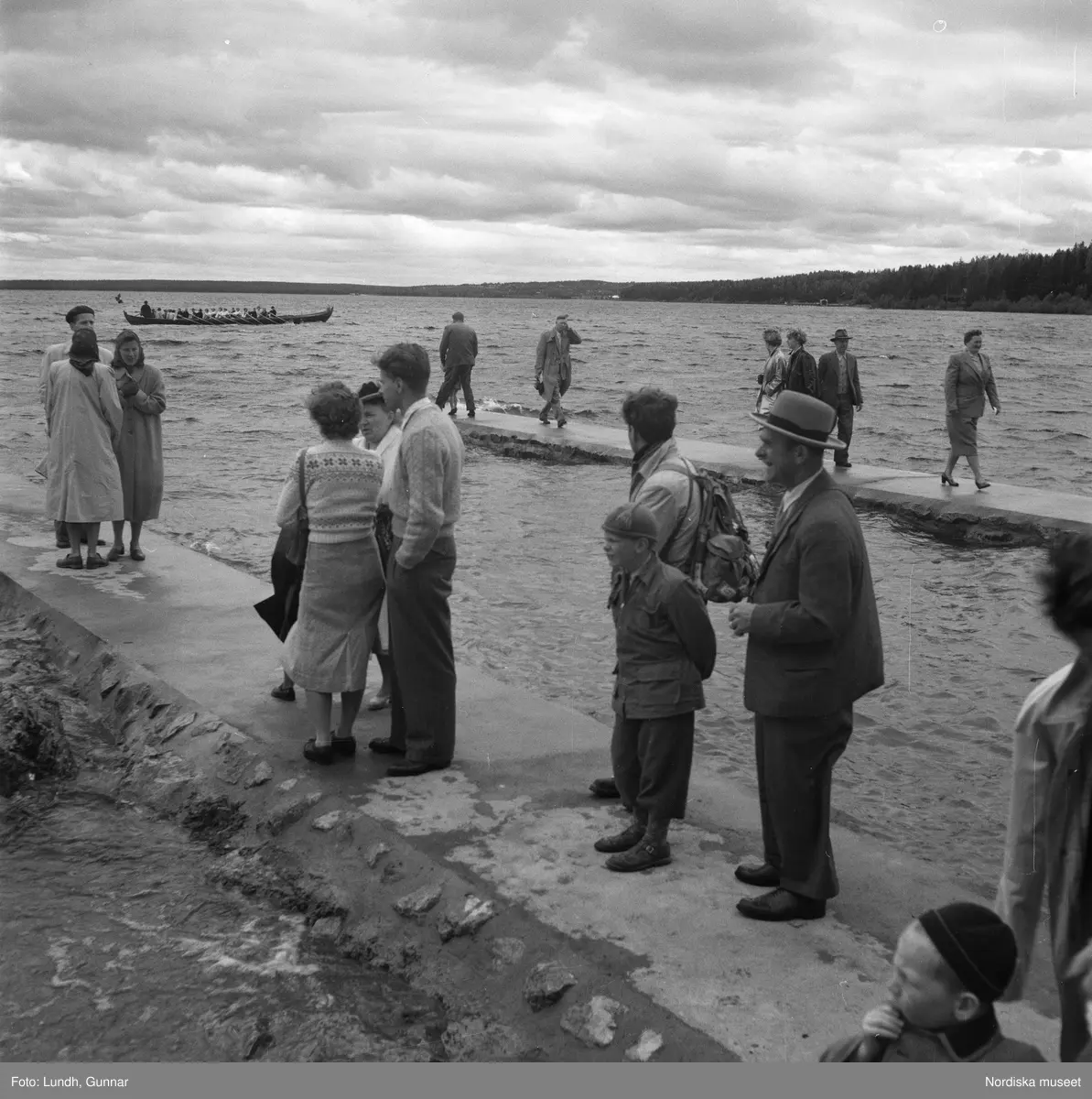 Motiv: (ingen anteckning) ;
En folksamling står vid en brygga med två kyrkbåtar, kvinnor och män ror kyrkbåtar i samband med midsommarfirande.