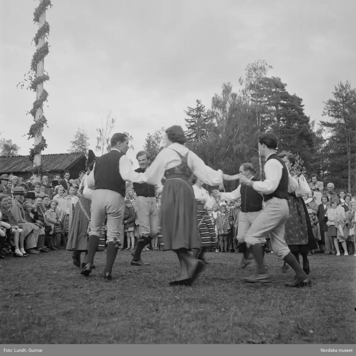 Motiv: (ingen anteckning) ;
Kvinnor och män i folkdräkt dansar vid en midsommarstång inför en publik vid midsommarfirande, tre hästskulpturer 2Jemt-Olofs hästar" (träsnidaren Jemt Olov Persson), porträtt av en man som står vid en husvägg, porträtt av en kvinna som står på en balkong.