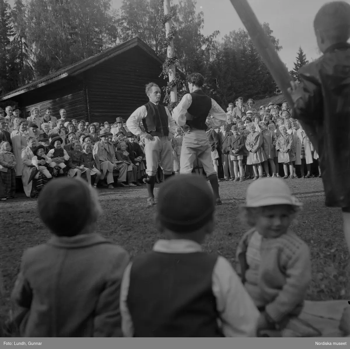 Motiv: (ingen anteckning) ;
Kvinnor och män i folkdräkt dansar vid en midsommarstång inför en publik vid midsommarfirande, tre hästskulpturer 2Jemt-Olofs hästar" (träsnidaren Jemt Olov Persson), porträtt av en man som står vid en husvägg, porträtt av en kvinna som står på en balkong.