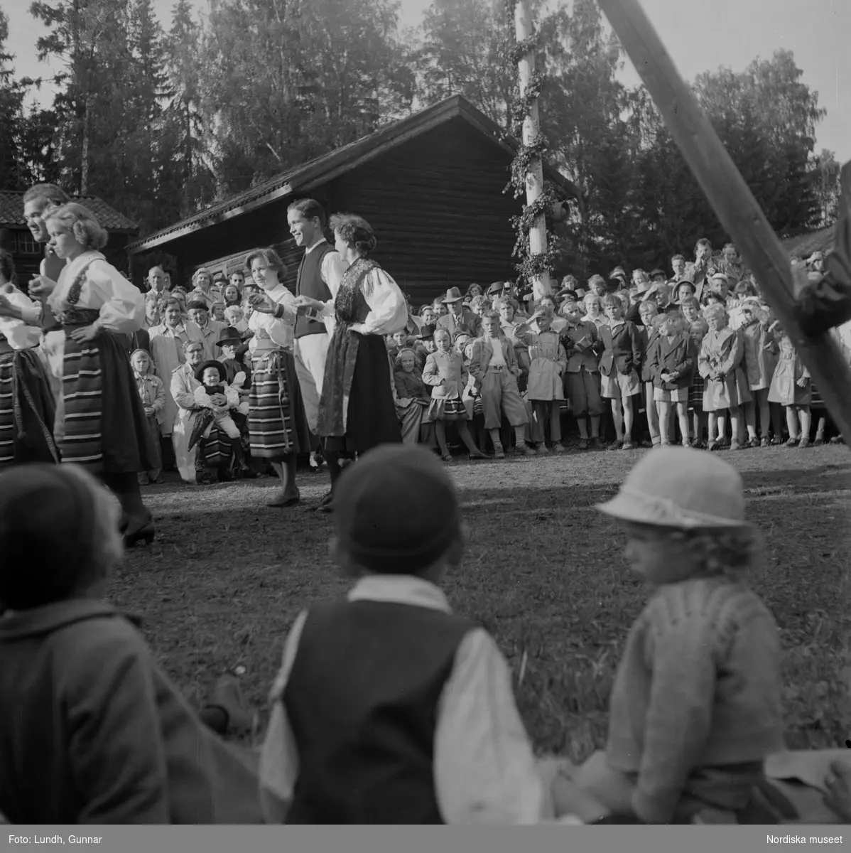 Motiv: (ingen anteckning) ;
Kvinnor och män i folkdräkt dansar vid en midsommarstång inför en publik vid midsommarfirande, tre hästskulpturer 2Jemt-Olofs hästar" (träsnidaren Jemt Olov Persson), porträtt av en man som står vid en husvägg, porträtt av en kvinna som står på en balkong.