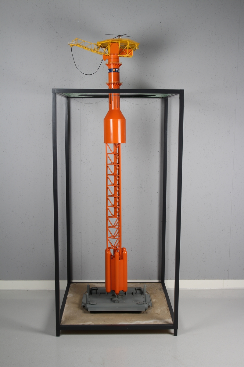 Modell av lastebøyen knyttet til Statfjord A. Malt orange og gul med helikopter på helidekket. Modellen holdes på plass av en aluminiumsramme der bunn og topp forestiller hhv. havbunn og hav.