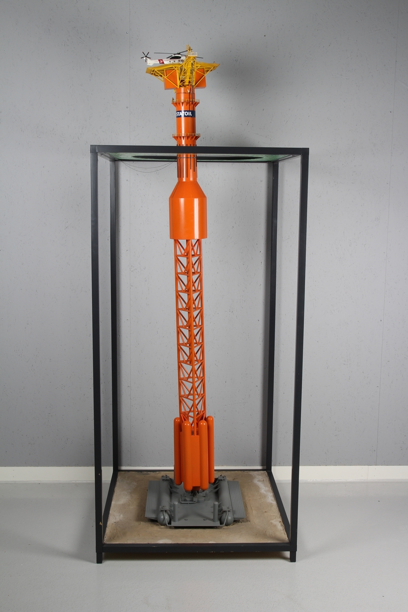 Modell av lastebøyen knyttet til Statfjord A. Malt orange og gul med helikopter på helidekket. Modellen holdes på plass av en aluminiumsramme der bunn og topp forestiller hhv. havbunn og hav.
