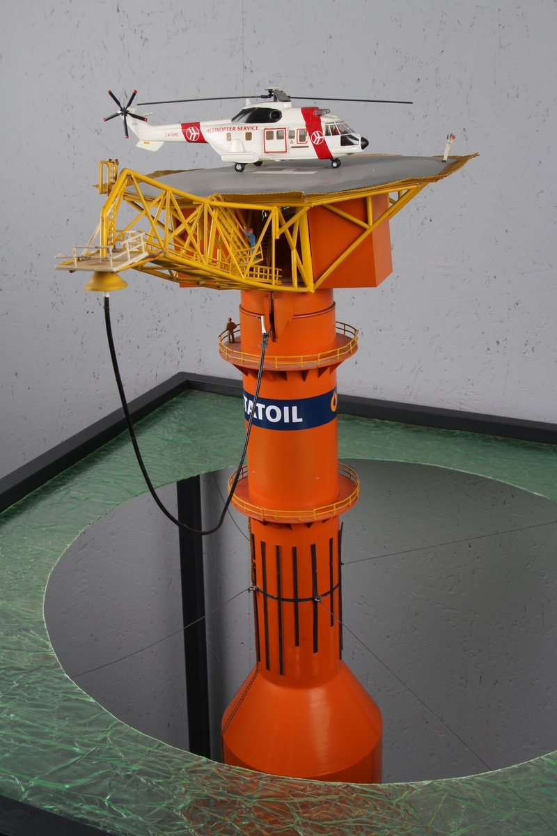 Modell av lastebøyen knyttet til Statfjord A. Malt orange og gul med helikopter på helidekket. Modellen holdes på plass av en aluminiumsramme der bunn og topp forestiller hhv. havbunn og hav.