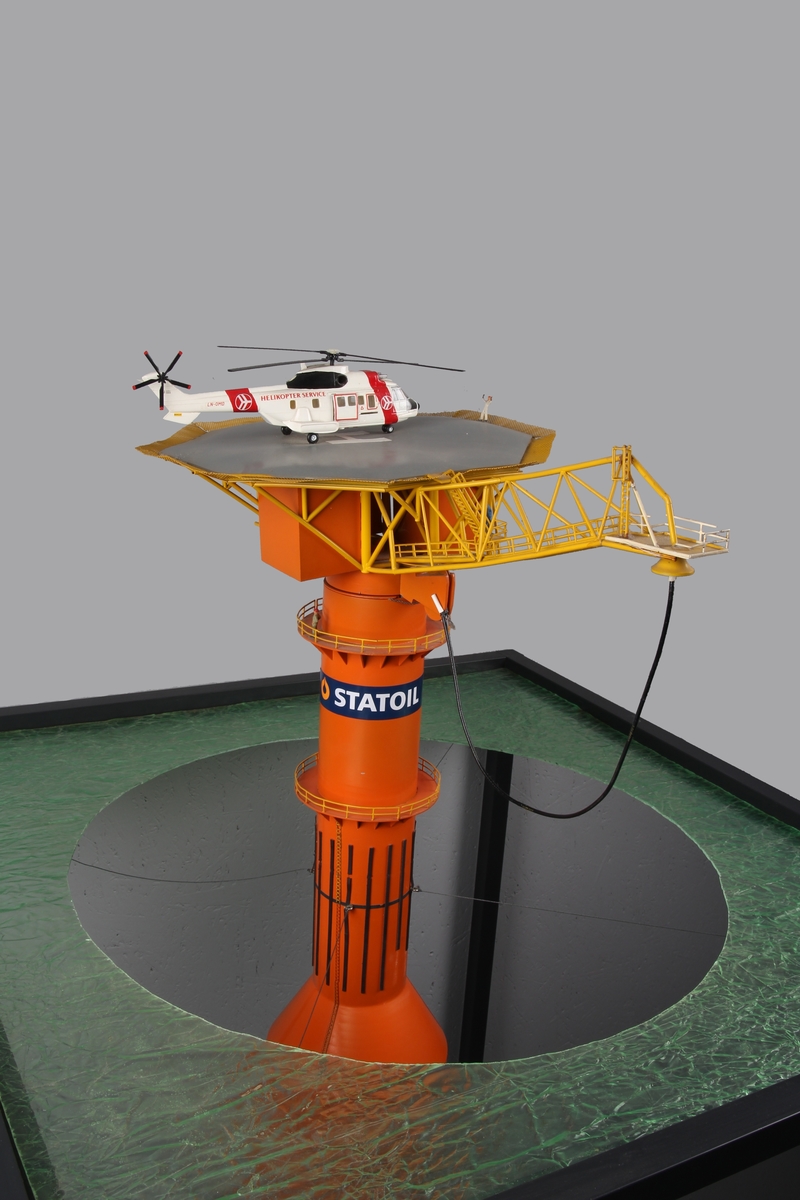 Modell av lastebøyen knyttet til Statfjord A. Malt orange og gul med helikopter på helidekket. Modellen holdes på plass av en aluminiumsramme der bunn og topp forestiller hhv. havbunn og hav.