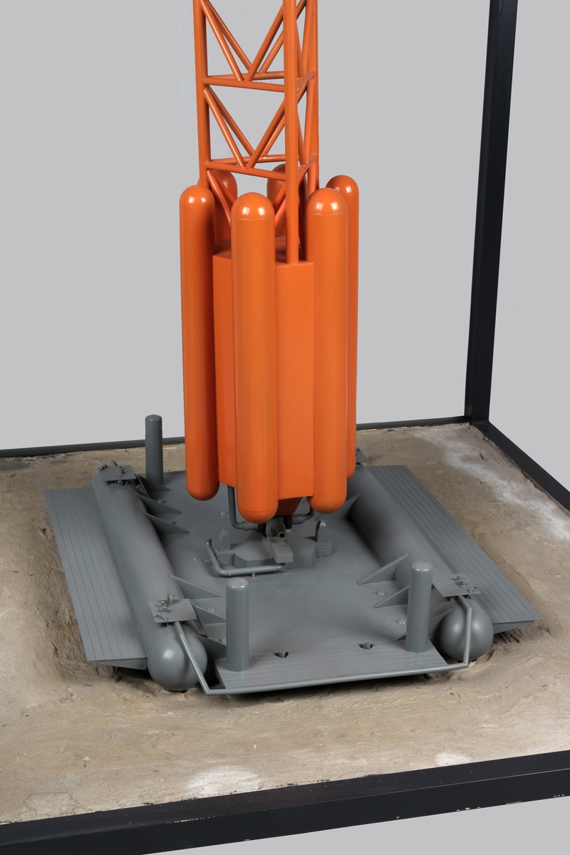 Modell av lastebøyen knyttet til Statfjord A. Malt orange og gul med helikopter på helidekket. Modellen holdes på plass av en aluminiumsramme der bunn og topp forestiller hhv. havbunn og hav.