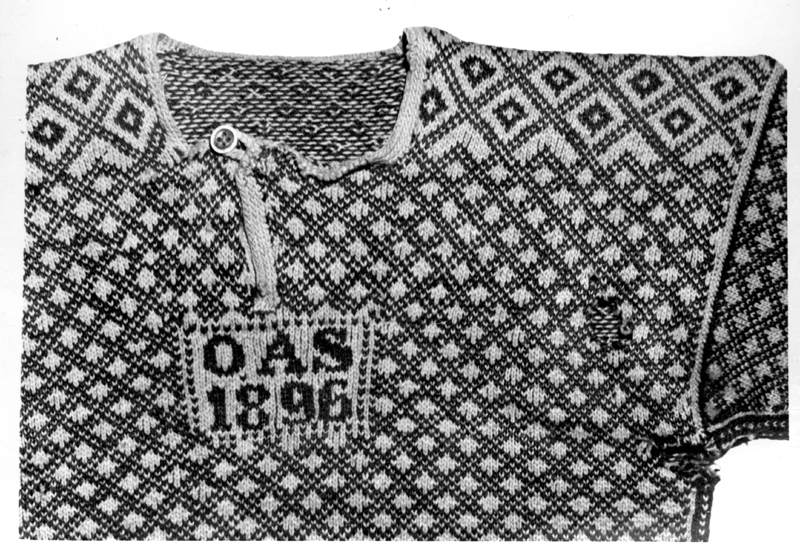 Stickad tröja av ylle. Röd med svart rutmönster, knäppt med en knapp i halsen. På bröstet instickat "O A S 1896" 
Nedtill en 5 cm bred bård. Längd: 58 cm, axelbredd: 56 cm, ärmlängd: 45 cm.